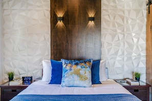 A modern hotel room with a geometric-patterned wall, a wooden headboard, and a bed with blue and white bedding. There are two bedside tables with lamps and a digital clock. - Luxurious STUDIO 19 - Quiet - Views  - Town in 5 mins - JTNP - in  8 minutes (Joshua Tree)