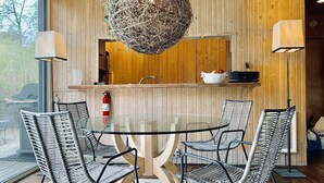 A modern dining area with a glass table surrounded by wicker chairs, a wooden wall with a hanging spherical light fixture, and a wooden counter with a sink.