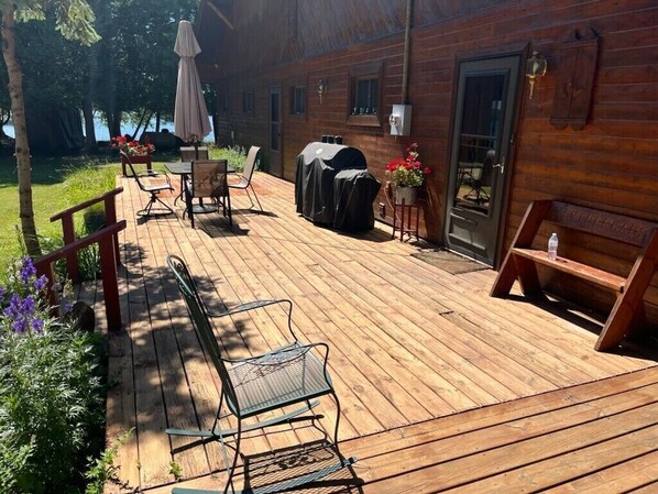 Una terraza de madera con mobiliario exterior, una parrilla y un pequeño jardín con flores.