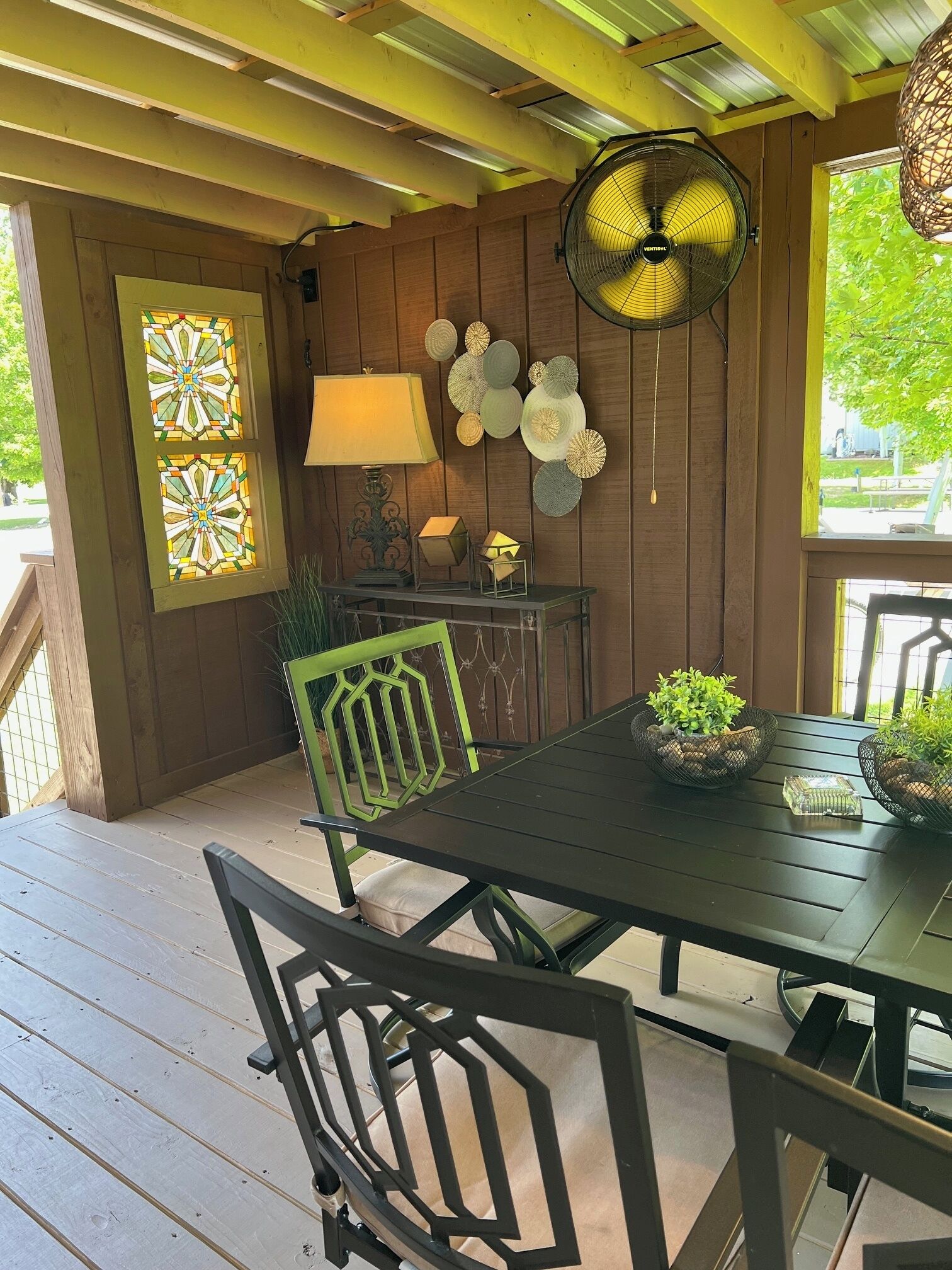 A wooden deck with a table and chairs, a fan, a lamp, and decorative wall art.