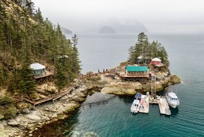 Una cabaña costera con terraza de madera, un muelle pequeño para barcos y un bosque en el fondo.