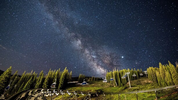 Cielo estrellado con la Vía Láctea visible, un telesilla y una cabaña pequeña en un área boscosa.