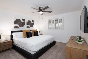 Un dormitorio con cama, mesita de noche, escritorio, ventilador de techo y ventana con contraventanas.