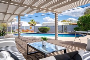 Zona de piscina con un porche techado, sillas de descanso y una mesa de vidrio.