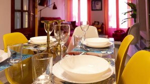 Una mesa con vajilla, vasos y cubiertos, rodeada de sillas amarillas en una habitación con una puerta roja y ventanas.