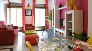 Una sala de estar con un sofá rojo, una mesa de centro de vidrio y una silla amarilla. Hay un televisor sobre un soporte blanco y una planta en el alféizar de la ventana.