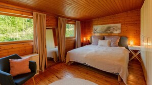 A cozy wooden cabin bedroom with a large bed, a chair, and a window offering a view of the forest.