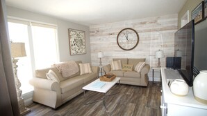 A living room with a beige sofa, a coffee table, a clock, and a television. - Cozy Condo at Sandpiper Cove/  pool and golf view (Destin)