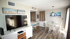 A compact living space with a flat-screen TV, a dining area with a table and chairs, and a kitchenette. - Cozy Condo at Sandpiper Cove/  pool and golf view (Destin)