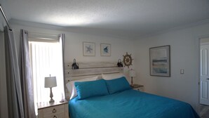 A bedroom with a bed, a nightstand, a lamp, a window with curtains, and wall decorations including a ship's wheel and framed pictures. - Cozy Condo at Sandpiper Cove/  pool and golf view (Destin)