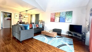 A modern living room with a grey sofa, a coffee table, and a flat-screen TV. There are abstract paintings on the wall and a wooden floor. - Executive Condo - Gigabit Wi-Fi - Near Redstone Arsenal - 2 beds-2 baths (Madison)