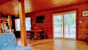 A wooden cabin interior with a patterned sofa, a dining table with chairs, and a large sliding glass door leading outside. - Lakefront Muskoka Beautiful Cottage  (Gravenhurst)