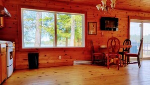 A cabin with a large window, wooden walls, a dining table with chairs, a TV mounted on the wall, and a fireplace. - Lakefront Muskoka Beautiful Cottage  (Gravenhurst)