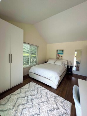 A bedroom with a bed, a wardrobe, a window with blinds, a painting on the wall, and a mirror.