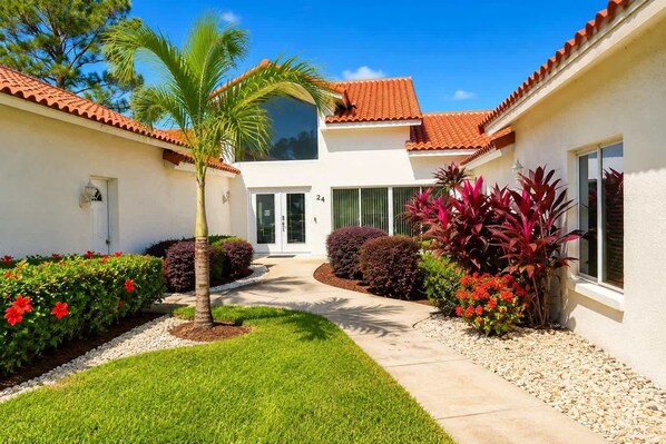A residential building with a tiled roof, a central entrance, and a well-maintained garden with various plants and a palm tree.