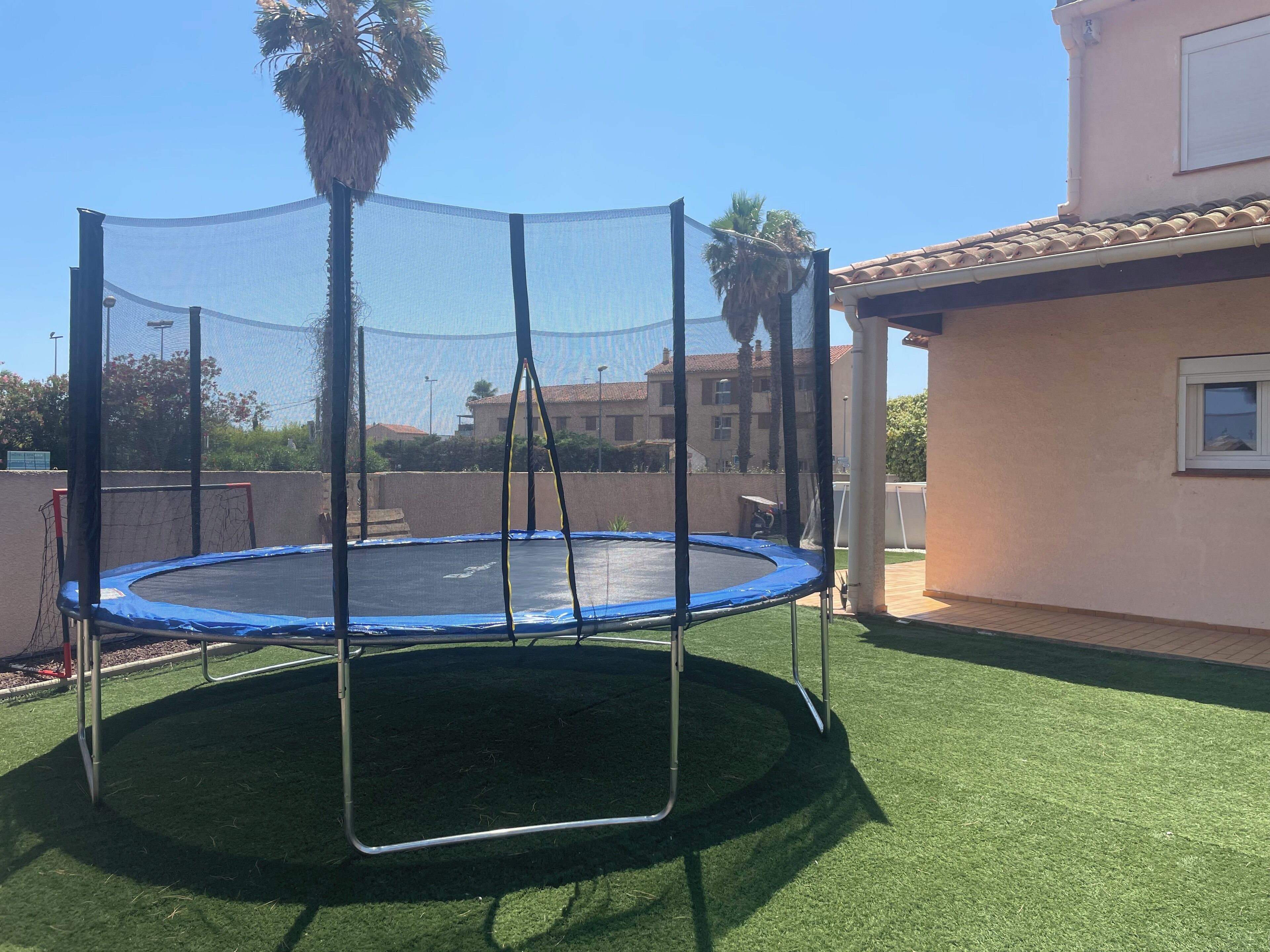 Een trampoline met veiligheidsnet, omringd door kunstgras, met een huis op de achtergrond.