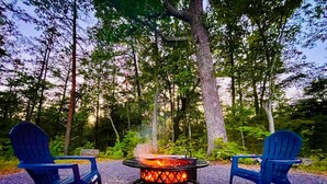 Two Adirondack chairs facing a circular fire pit with a visible flame, set on a gravel path surrounded by trees. - Romantic Cabin, Mnt View, Perfect Location❤️ (Gatlinburg)