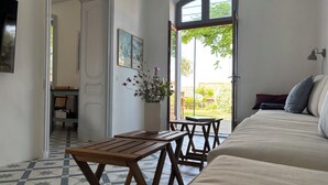 A room with a bed, a wooden table, a vase with flowers, and a view of the garden through a large window.