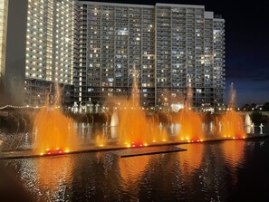 Vue nocturne d’un hôtel à plusieurs étages, doté de balcons, illuminé par des lumières provenant de l’intérieur. Au premier plan, on aperçoit une fontaine avec des jets d’eau et des jeux de lumière.