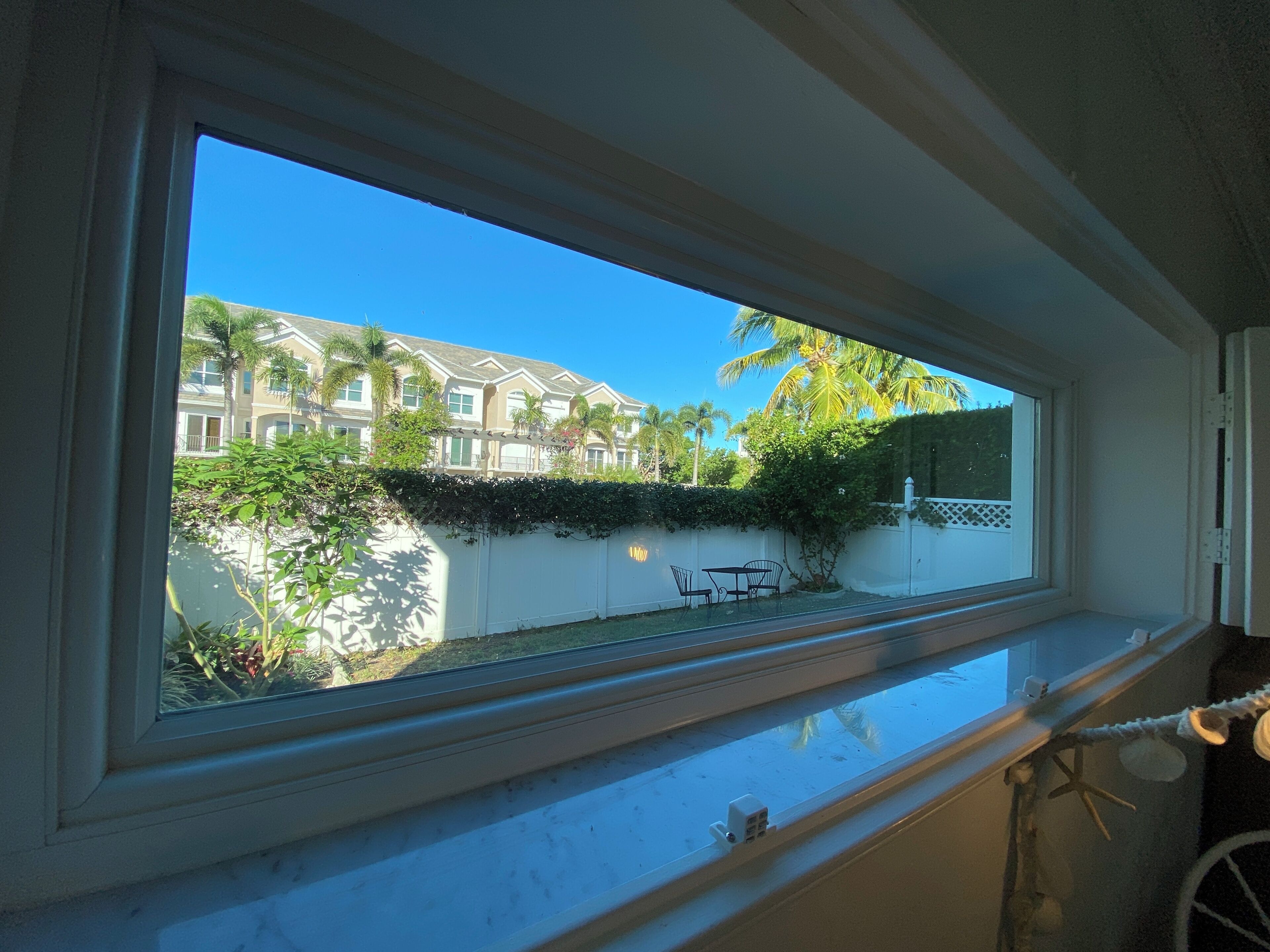 A window with a view of a residential area, including a white fence, a palm tree, and a building with multiple windows.