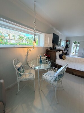 Quarto com mesa de vidro redonda, cadeiras brancas, cama, televisão e janela com vista para casas e áreas verdes.