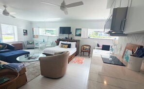 Uma sala de estar moderna com sofá, poltrona e uma cama com travesseiro 'Relax'. Há ventiladores no teto, uma televisão e uma cozinha com micro-ondas.