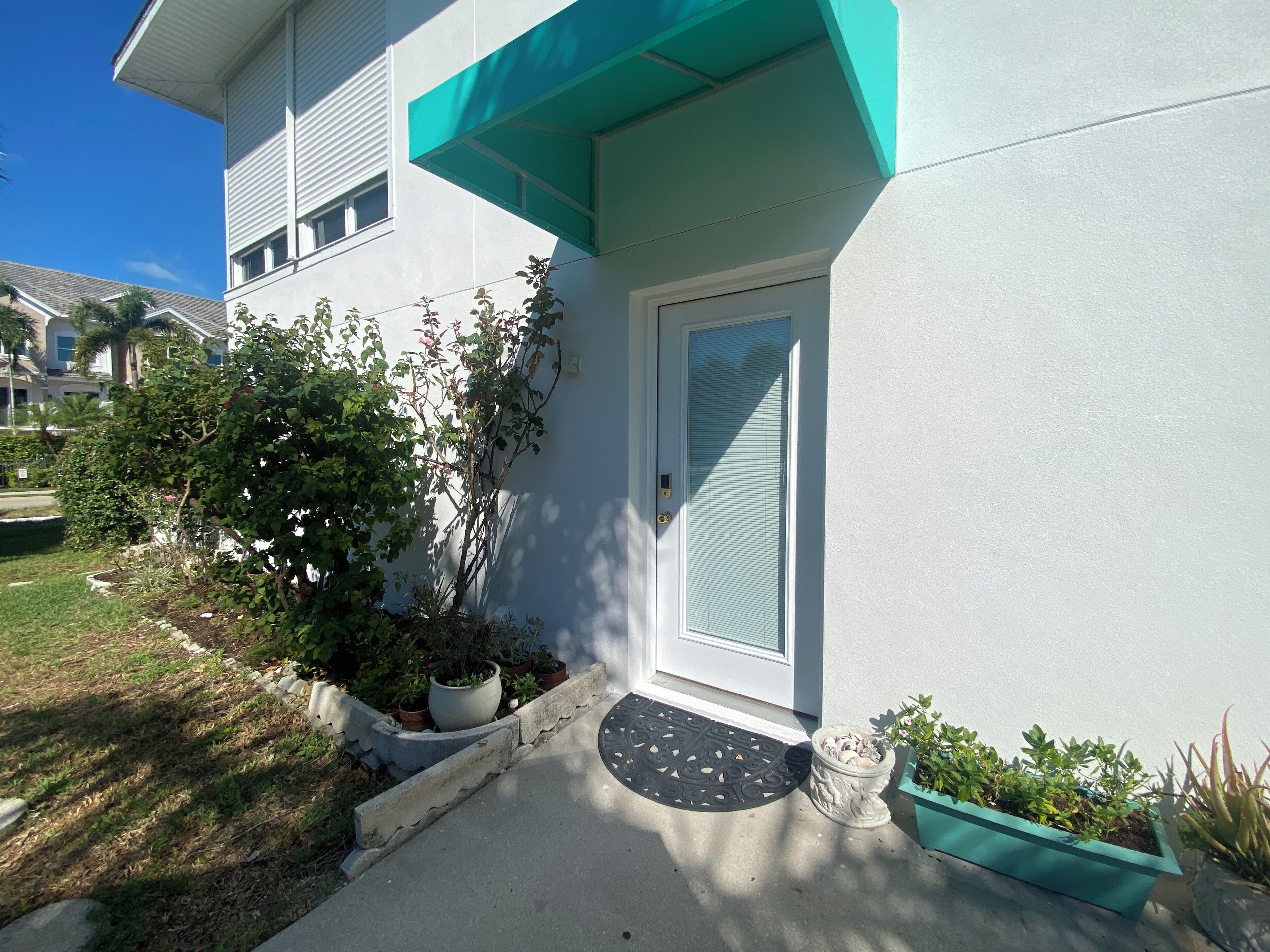 A modern house with a white exterior, a blue awning, and a front door with a glass panel. There are potted plants and a doormat on the front porch.