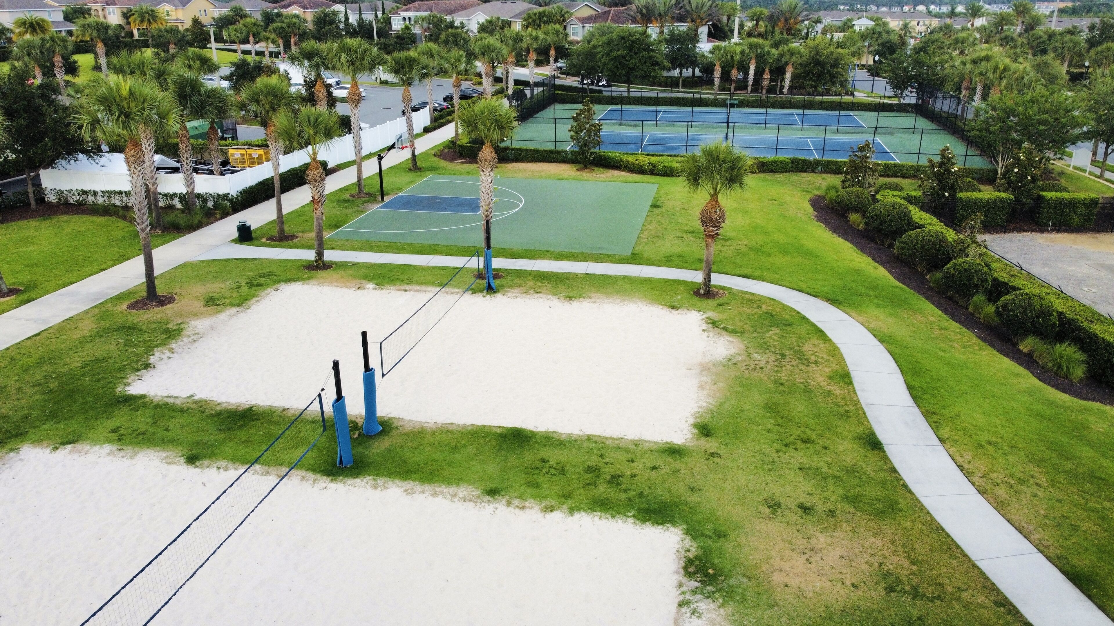 An aerial view of a tennis court surrounded by palm trees and a walking path.