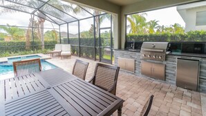 Une terrasse extérieure avec une piscine, une table en bois, des chaises et une cuisine attenante équipée d’un barbecue.