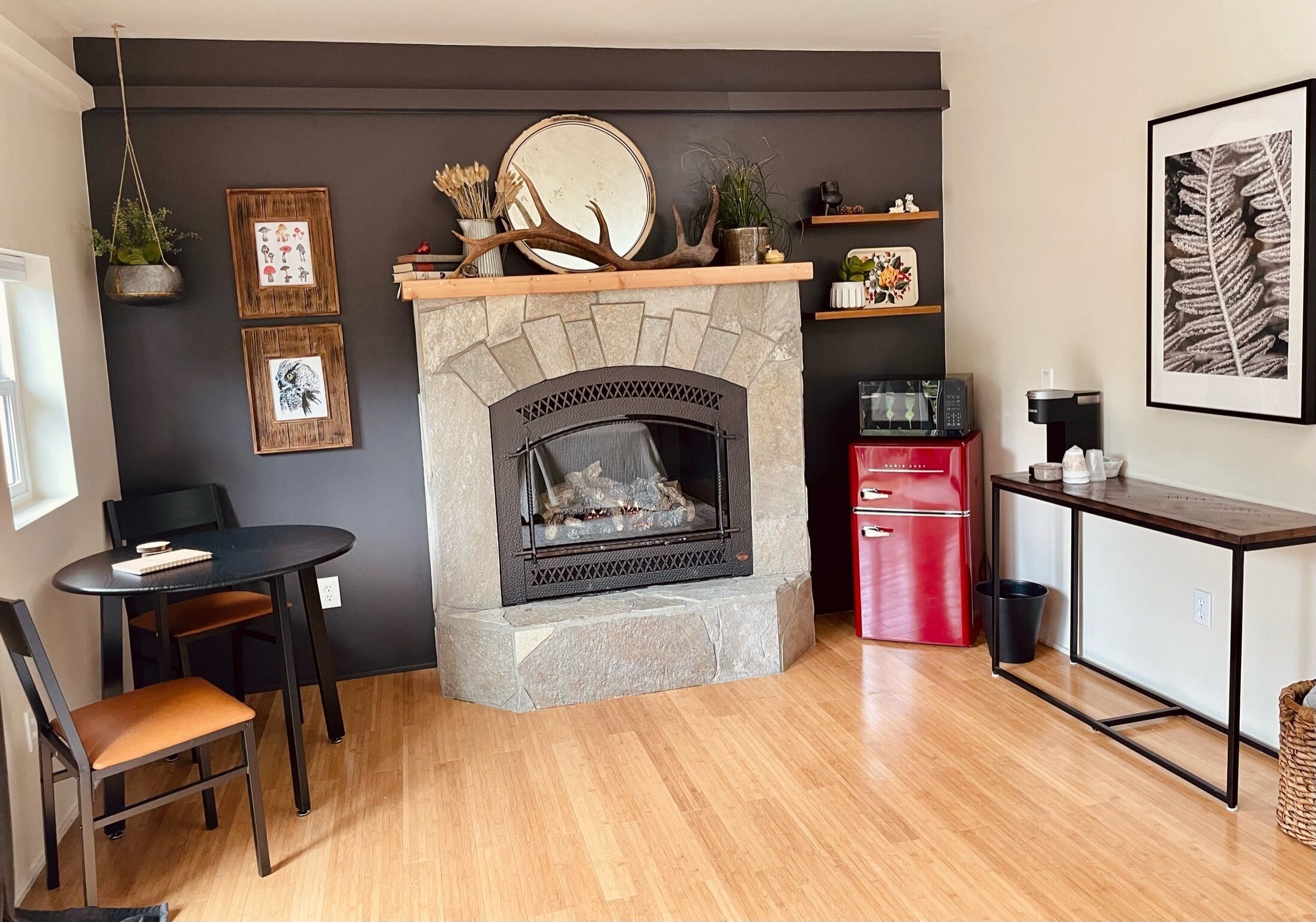 A modern living room with a stone fireplace, wooden shelves, a dining table with chairs, and a red appliance.