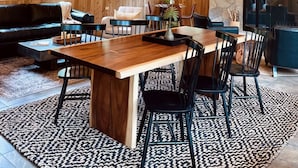 Una habitación con paneles de madera, una gran mesa de comedor, sillas negras y una araña. Hay dos ventanas y una chimenea de piedra.