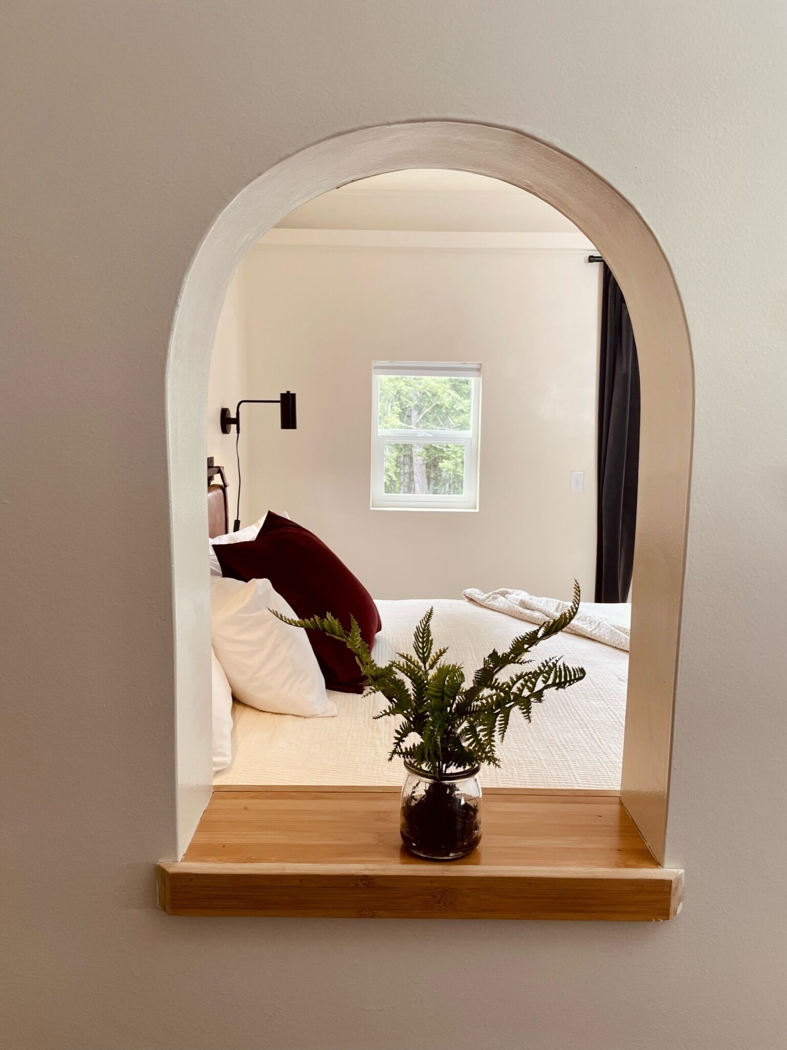 Un dormitorio con cama, ventana, lámpara de noche y una planta en un estante.