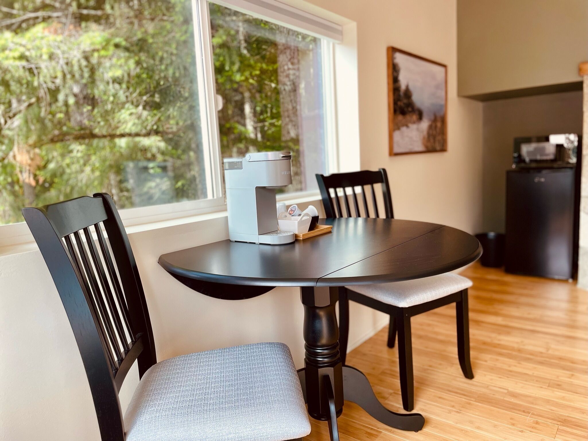 A dining area with a round table, two chairs, a coffee maker, and a refrigerator in the background.