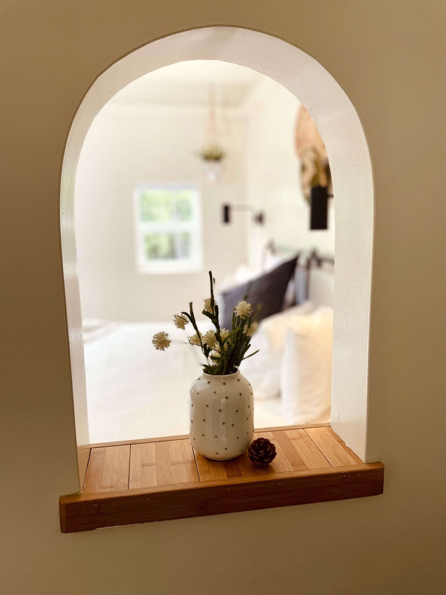 A small, white, polka-dotted vase with flowers on a wooden shelf, viewed through an arched window.