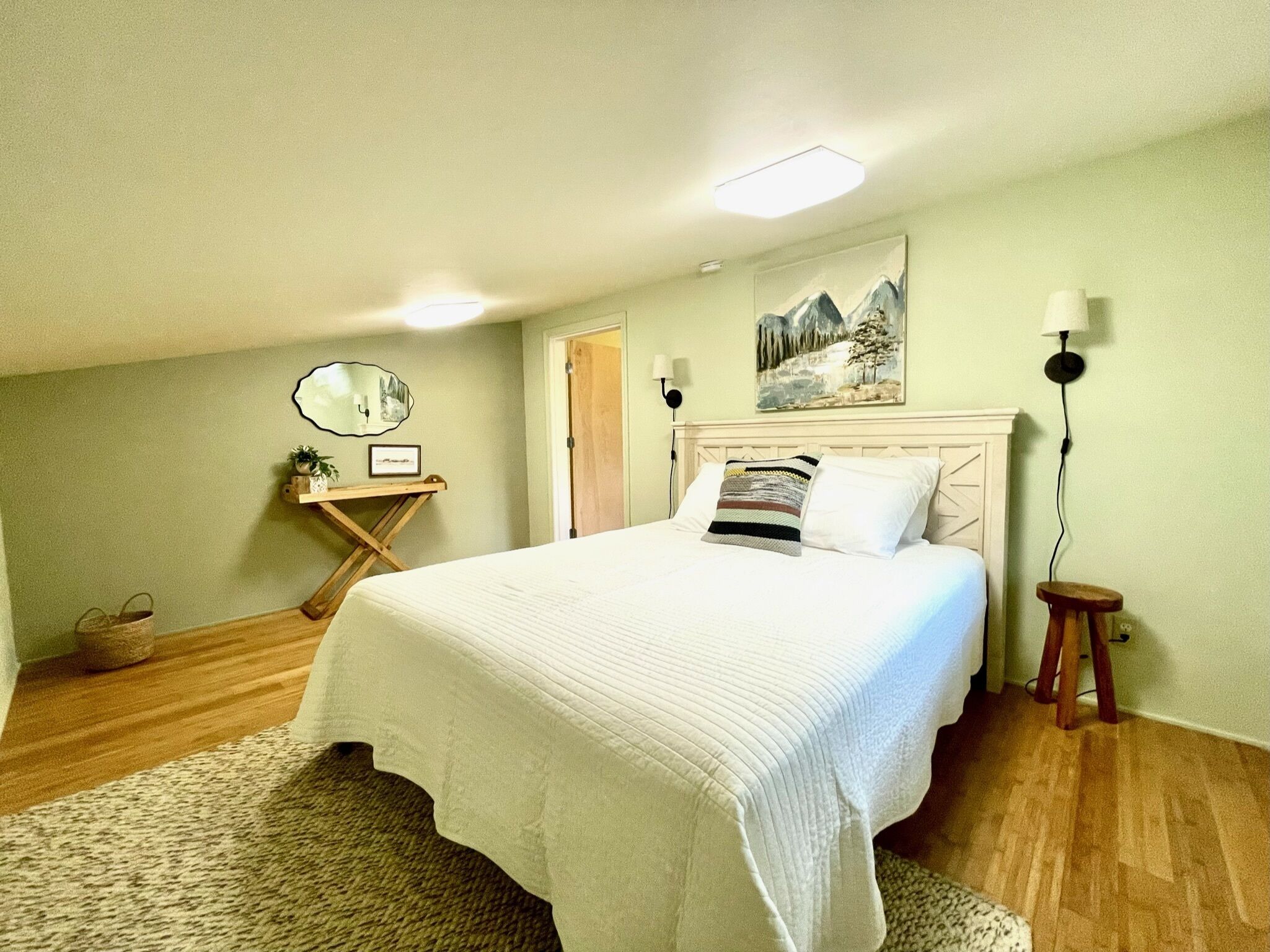 Un dormitorio con una cama grande, piso de madera, un cuadro en la pared y una mesita de noche con una lámpara.
