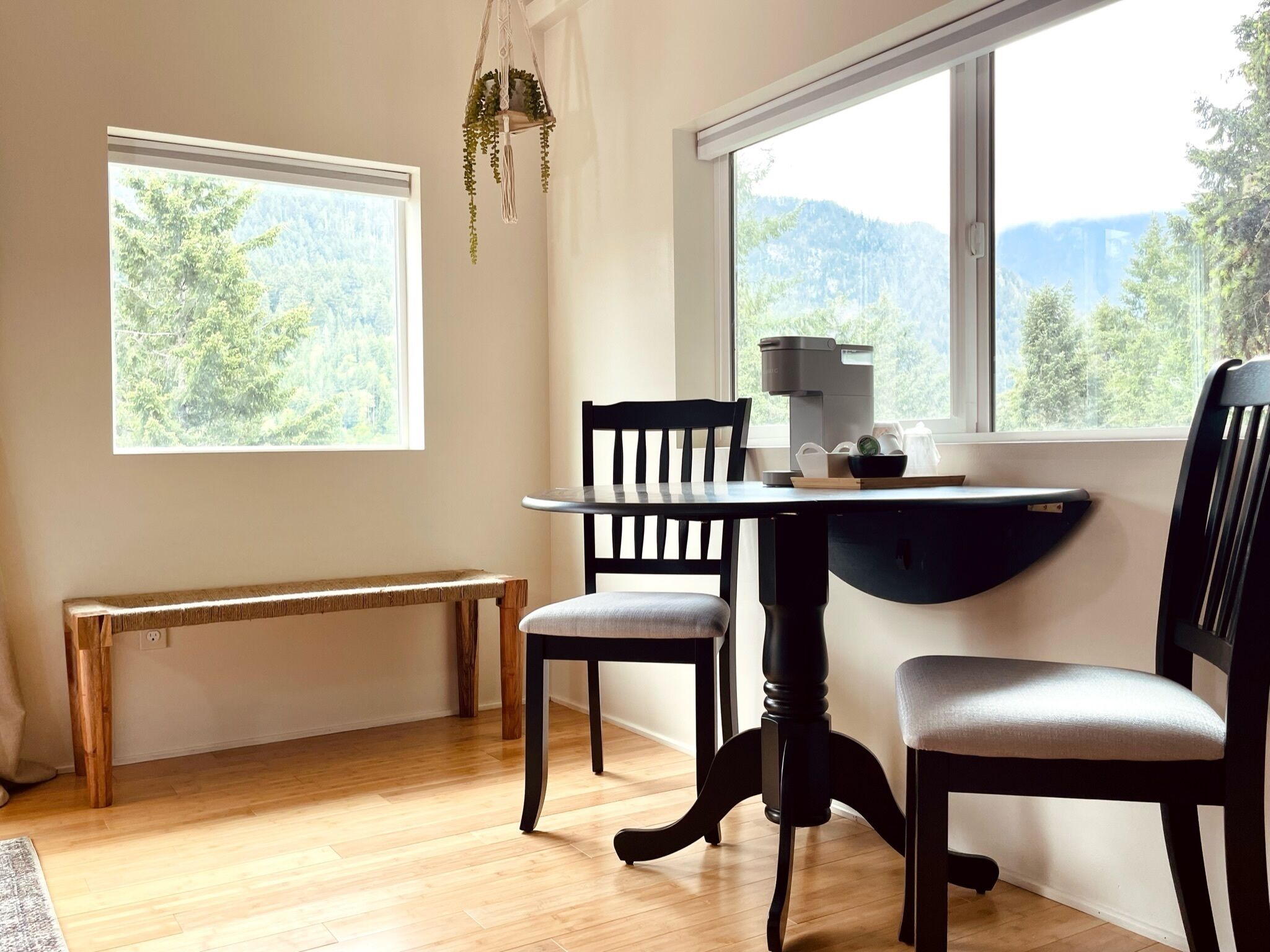 Une pièce avec un plancher en bois, une table ronde entourée de chaises, un banc et une fenêtre donnant sur des arbres et des montagnes.