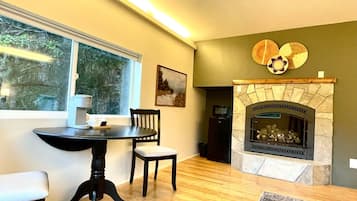 Een gezellige woonkamer met een open haard, een eettafel met stoelen en een raam met uitzicht op bomen.