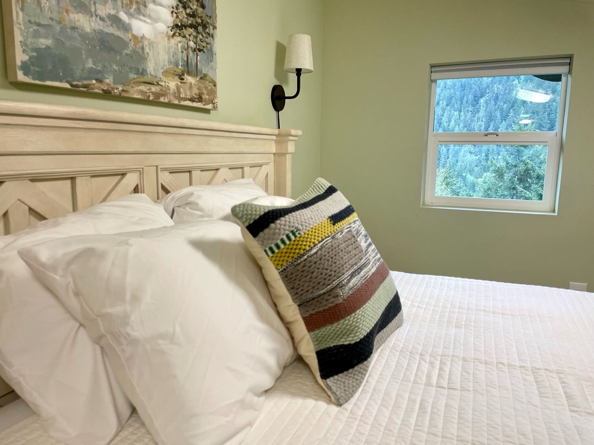 Ein Bett mit weißer Bettwäsche und einem farbenfrohen Streifenmuster-Kissen, ein Gemälde an der Wand, eine Wandlampe und ein Fenster mit Blick auf einen Wald.