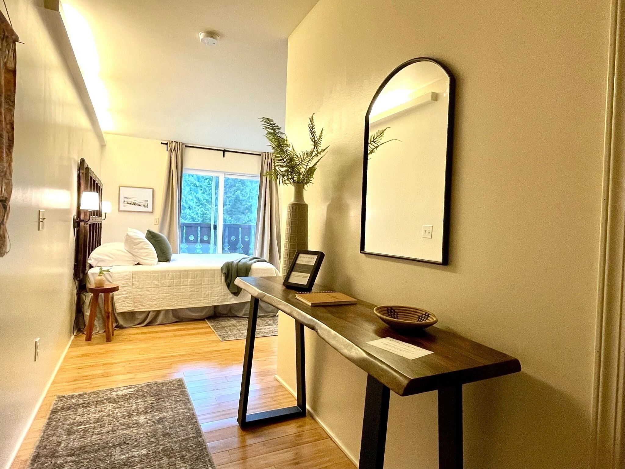A hotel room with a bed, a bedside table with a vase, a mirror, and a view of the outdoors.