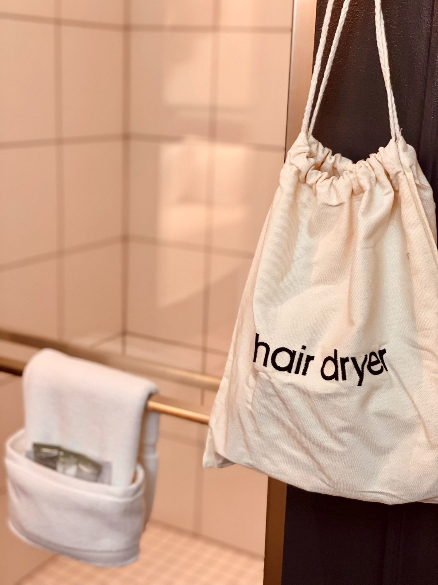 Une salle de bain avec un porte-serviettes sur lequel est posée une serviette blanche et un sac beige à cordon de serrage sur lequel est inscrit le mot « sèche-cheveux ».