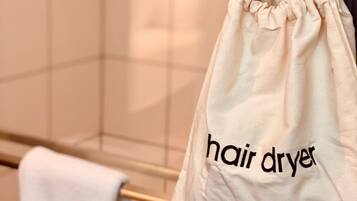 Une salle de bain avec un porte-serviettes sur lequel est posée une serviette blanche et un sac beige à cordon de serrage sur lequel est inscrit le mot « sèche-cheveux ».