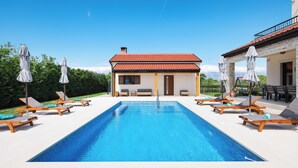 A swimming pool with lounge chairs and umbrellas, a house with a red-tiled roof, and a clear blue sky. - Villa Velebita (Zadar)