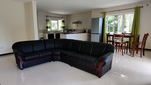 Ein modernes Wohnzimmer mit einem schwarzen Ecksofa, einem Essbereich mit Holzkreislern, einer Küche im Hintergrund und einem großen Fenster mit Vorhängen.