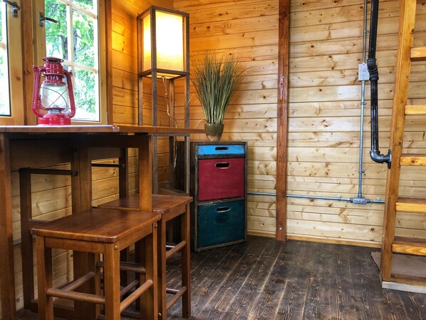Un interior de madera con una mesa de madera, dos taburetes, un farol, una lámpara de pared y un mueble bajo.