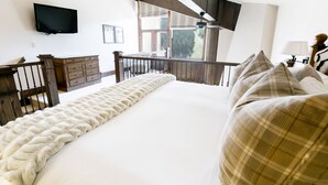 A bedroom with a large bed, a wooden dresser, a TV mounted on the wall, and a balcony with a view of trees.