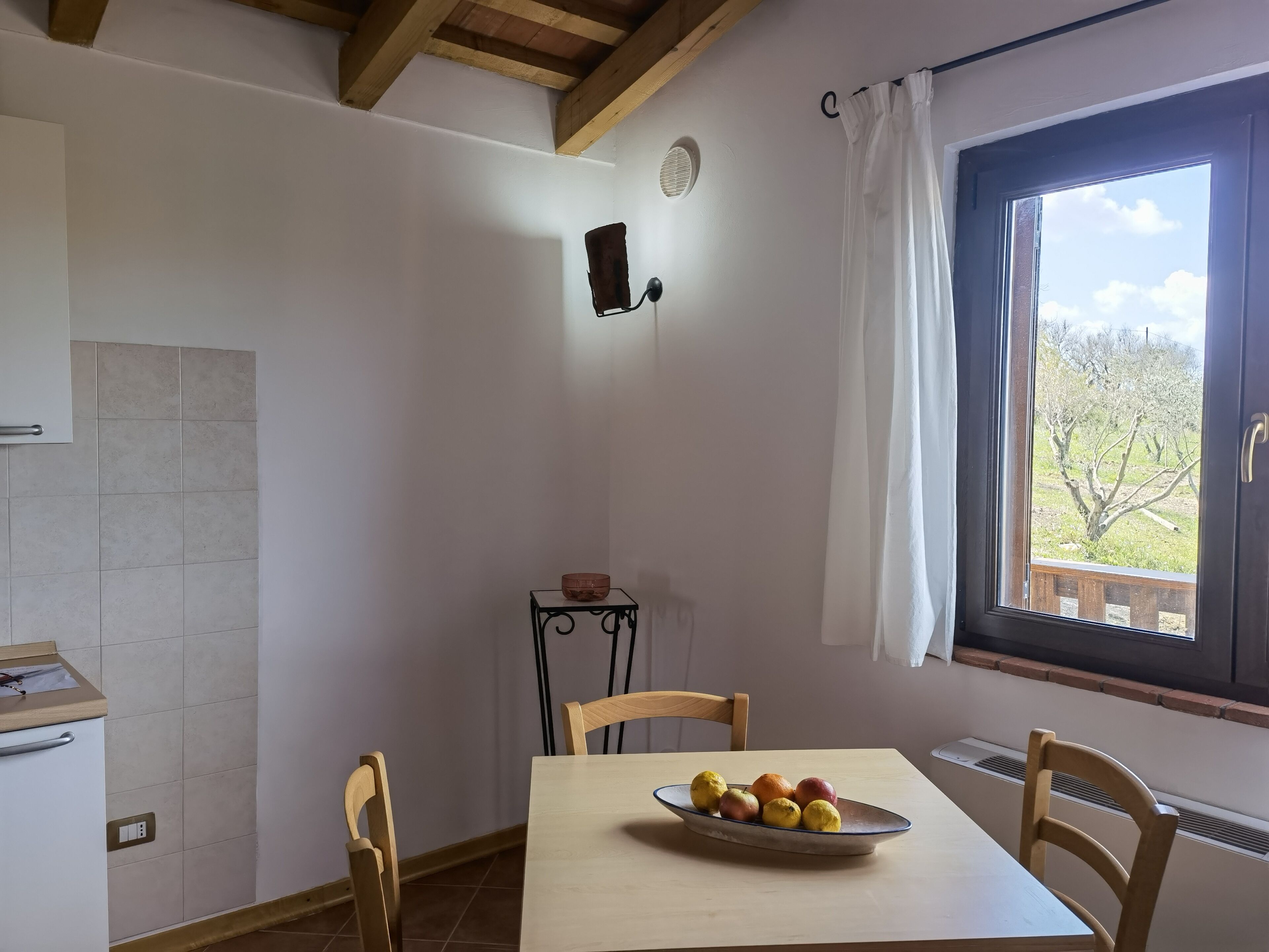 A room with a wooden table, chairs, a window with a view of trees, a radiator, and a small shelf with a bowl.