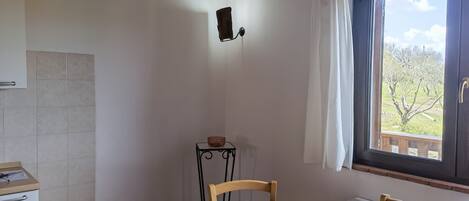 Una habitación con una mesa de madera, sillas, una ventana que da a árboles, un radiador y un estante con un cuenco.