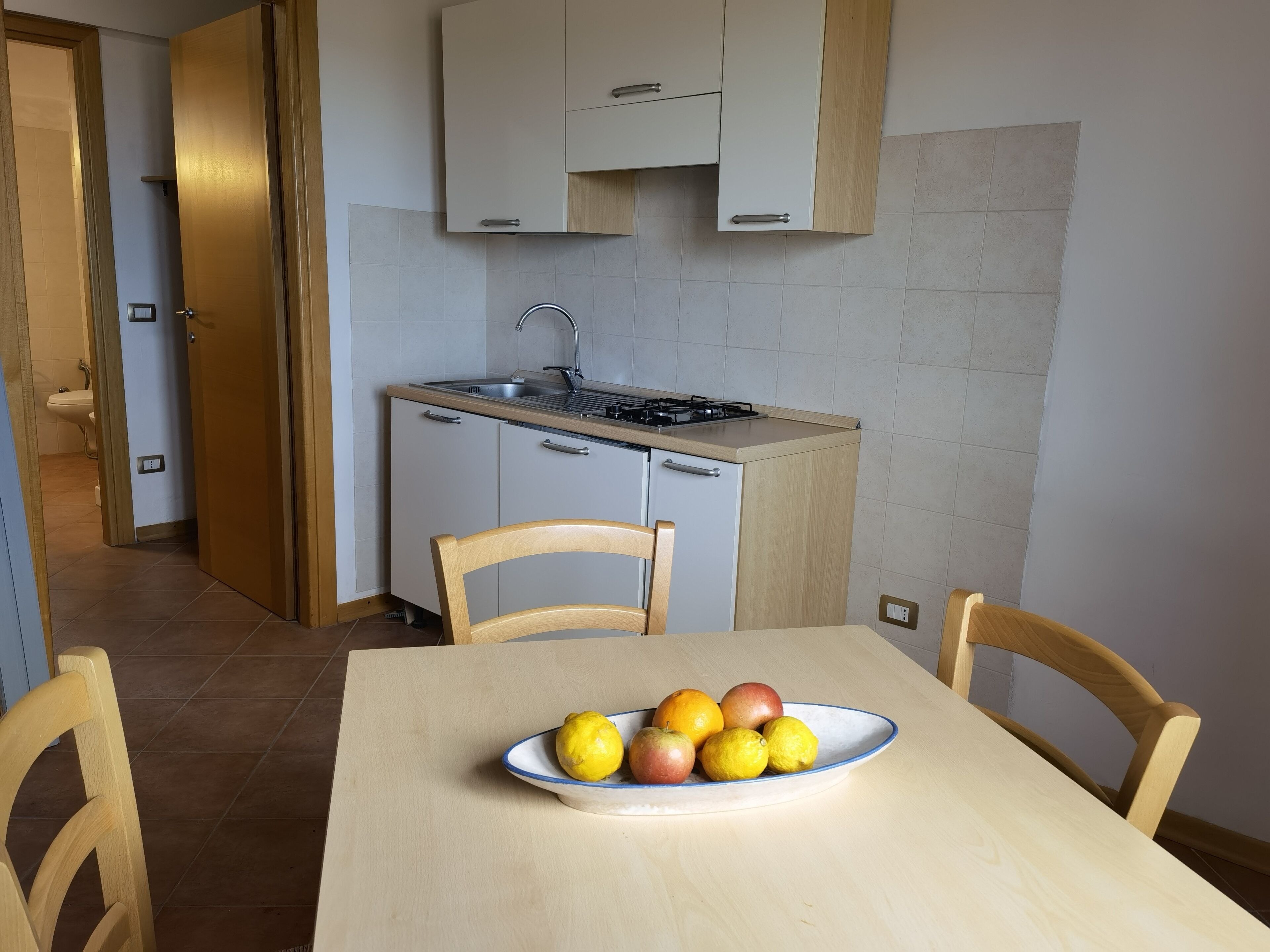 A compact kitchen with a sink, stove, and cabinets. A table with chairs and a bowl of fruit is in the foreground.
