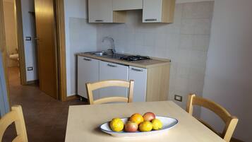A compact kitchen with a sink, stove, and cabinets. A table with chairs and a bowl of fruit is in the foreground.