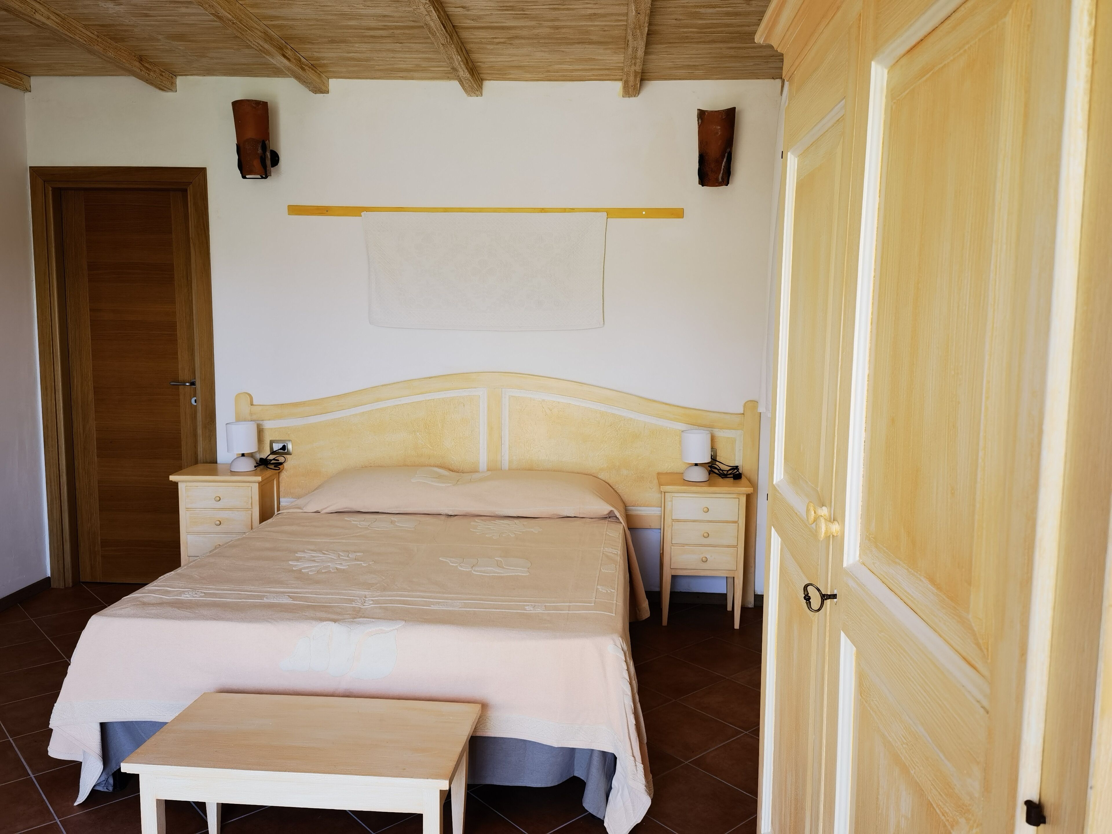 Ein Schlafzimmer mit einem hölzernen Bett, Nachttischen, einem Beistelltisch, einer Bank und einer Tür.